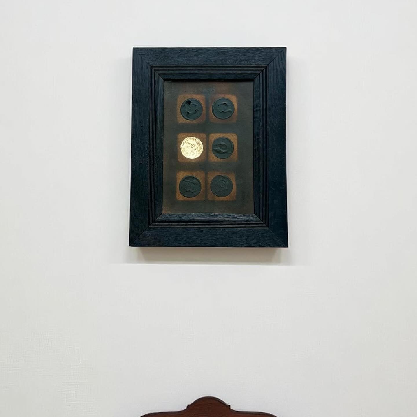 Framed artwork with six round objects on a white wall, with a wooden chair partially visible.